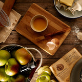 An overhead view of a table that has various things on it, such as a wooden chopping block, a whisk, a rolling pin, some juicy looking apples and a slab of butter