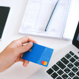 A close up of a hand holding a credit card. Also in the picture is a laptop, a notepad and pencil and a mobile phone