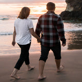 A man and a woman are walking along a beach, holding hands. The sun is just setting and a wave can be seen lapping onto the sand