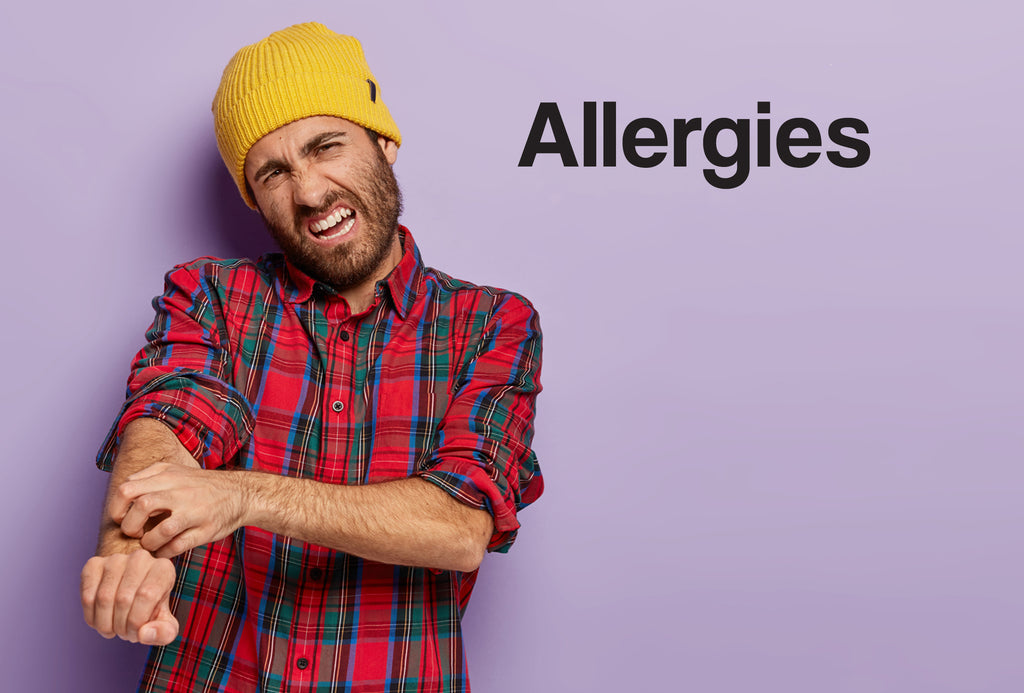 A man in a red lumberjack shirt and a yellow beanie is scratching his arm – the man looks in pain. The word – Allergies – can also be seen