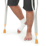 The lower half of a man; his right foot is bandaged and his left foot is without shoes and socks. There are two crutches that the man is using that are on either side of him