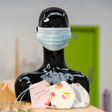 An image of a manequin with a blue surgical mask over its mouth, with 3 floral, hand made masks in front of it