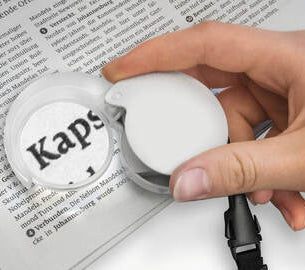 A Eschenbach Mobilent Compact Magnifier being held over some newsprint. The type in the magnifier is enlarged
