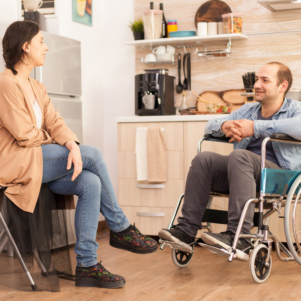 Set in a kitchen, a man sits in a wheelchair, while a woman is sitting on a chair. The two are engaged in conversation