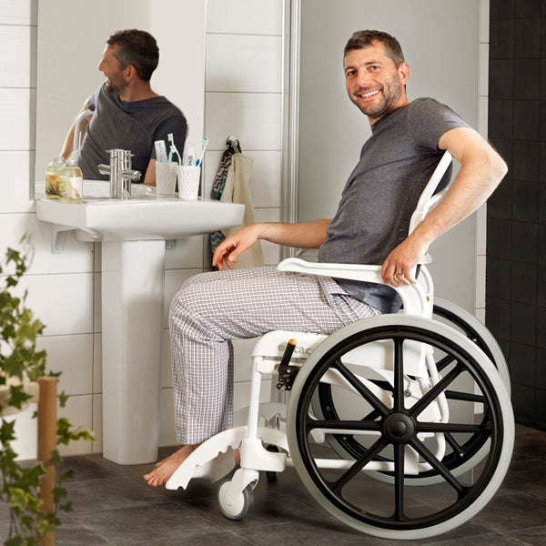 The image shows a young man using the Etac Clean Self Propelled Shower Commode Chair to help get him to the bathroom sink