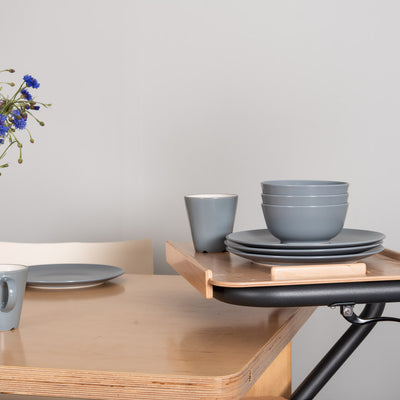 The image shows the SALJOL Beech Tray for Page Indoor Rollator, with a stack of plates and bowls and a cup on top of it, stood alongside a kitchen table.