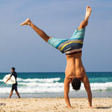 A picture showing a sandy beach with waves breaking on the shore. A man in trunks can be seen cartwheeling across the sand. There is a man in the distance is walking towards the sea carrying a surfboard