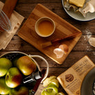 An overhead view of a table that has various things on it, such as a wooden chopping block, a whisk, a rolling pin, some juicy looking apples and a slab of butter