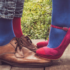 Some brown men's shoes with a men wearing some blue and pink socks and some pink, women's stilettos with the woman wearing blue socks