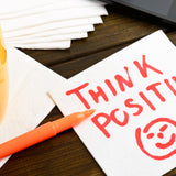 A table top with a yellow cup with black coffee inside on it. There is also a big note with the words – THINK POSITIVE – on it, with a scribble of a happy face
