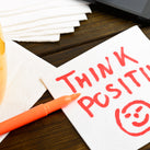 A table top with a yellow cup with black coffee inside on it. There is also a big note with the words – THINK POSITIVE – on it, with a scribble of a happy face