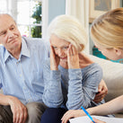 A picture of three people. The person in the middle is a lady who looks a little confused and is resting her head on both of her hands. A man sits to the left of her, looking concerned. A nurse sits to the right of her; she's holding a notepad