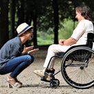 A lady in a wheelchair, in front of a group of trees, with another lady crouched in front of her – the pair are chatting