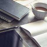 A picture of three notepads on a wooden table top. There is also a tablet, a cup of tea and a pencil also on the table