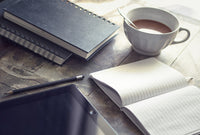 A picture of three notepads on a wooden table top. There is also a tablet, a cup of tea and a pencil also on the table