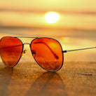 A pair orange lens sunglasses lying on some sand. The sun can be seen in the distance 