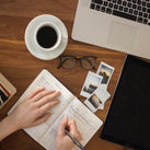 An overhead view of a table top, on which is a cup of coffee, a pair of glasses, a laptop and a diary, which someone is writing in