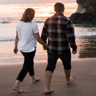 A man and a woman are walking along a beach, holding hands. The sun is just setting and a wave can be seen lapping onto the sand