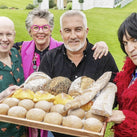 A photograph of all the hosts of The Great British Bake Off. They are holding a big wooden tray with a variety of baked goods.