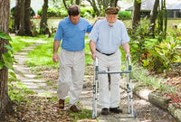 A man using a Zimmer frame on a path. He is being helped by a male friend