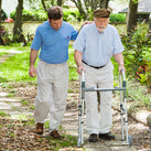 A man using a Zimmer frame on a path. He is being helped by a male friend