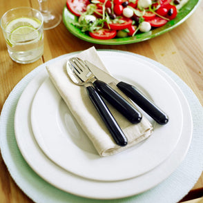 A single set of cutlery placed on top of a white dinner plate, with salad and drink located at the side.