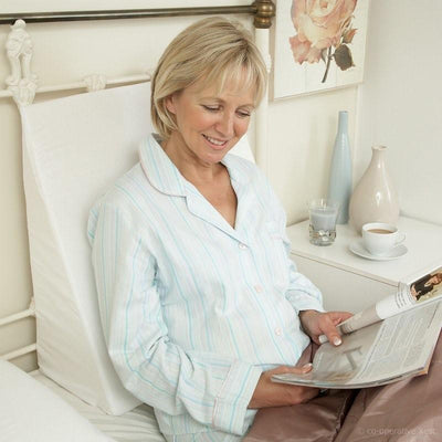 This image shows a middle aged lady in her pyjamas relaxing in her bed, using a harley bed relaxer whilst reading a magazine, she is smiling. 