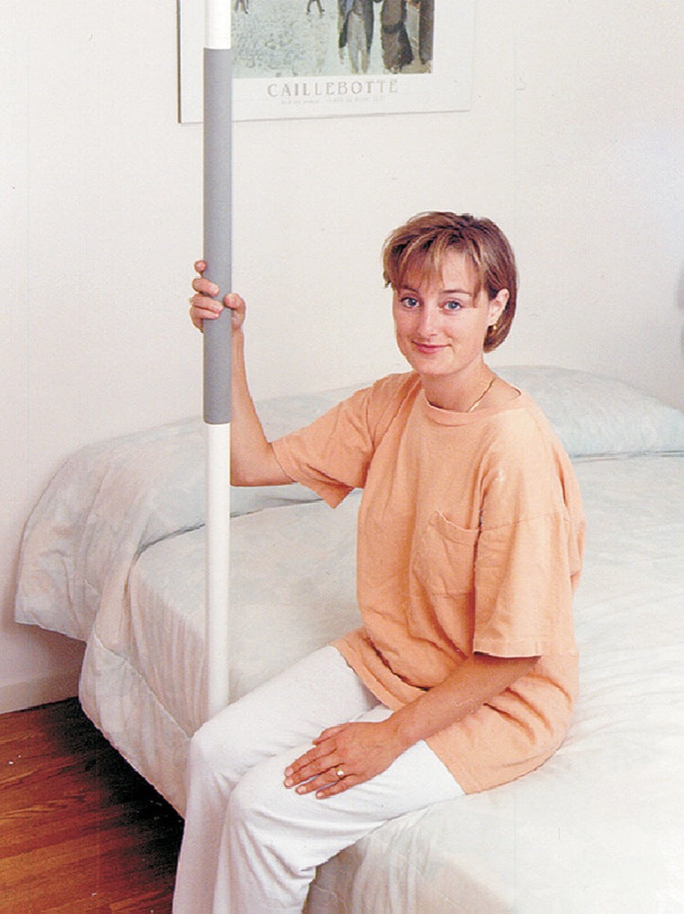 shows the  floor mounted SuperPole Grab Rail fitted beside a bed and a young woman sitting on the bed holding on to the rail.