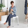 Image shows a woman sat on the edge shower stool in the bathroom. She is drying her hands near the sink.