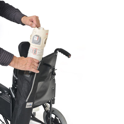 The image shows a man's hands inserting a wheelchair cylinder into the wheelchair oxygen bag which is fastened on to the back of a wheelchair