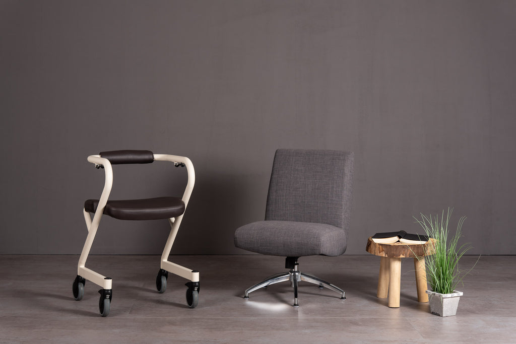 The image shows the Page Indoor Rollator with dark brown leatherette seat, alongside a chair and small coffee table