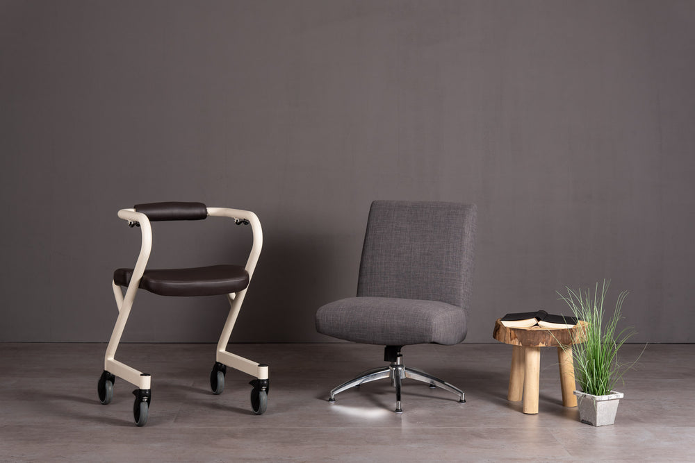 The image shows the Page Indoor Rollator with dark brown leatherette seat, alongside a chair and small coffee table
