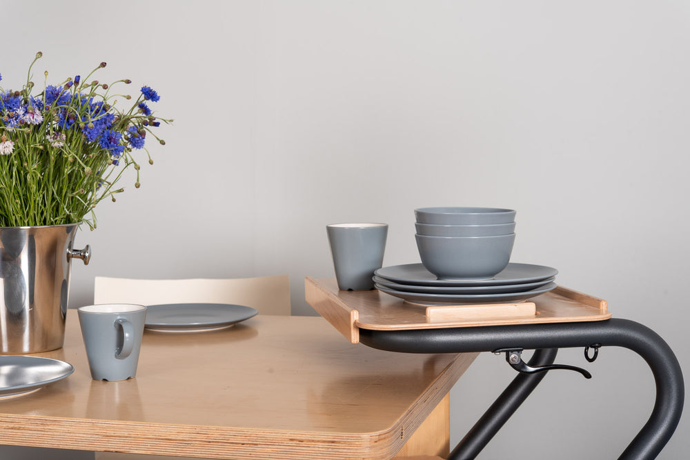 The image shows the SALJOL Beech Tray for Page Indoor Rollator, with a stack of plates and bowls and a cup on top of it, stood alongside a kitchen table.