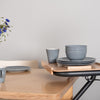 The image shows the SALJOL Beech Tray for Page Indoor Rollator, with a stack of plates and bowls and a cup on top of it, stood alongside a kitchen table.